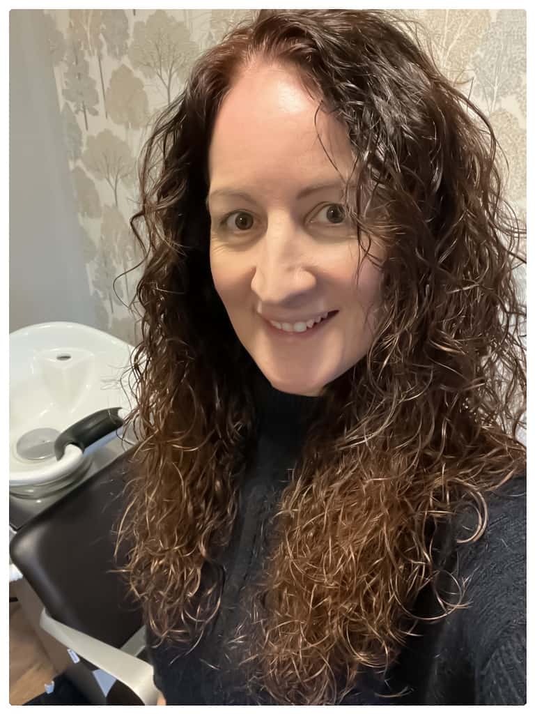 A photograph of Claire Gilbraith, standing in front of a sink at her salon. She is white with long curly dark hair and smiles at the camera.